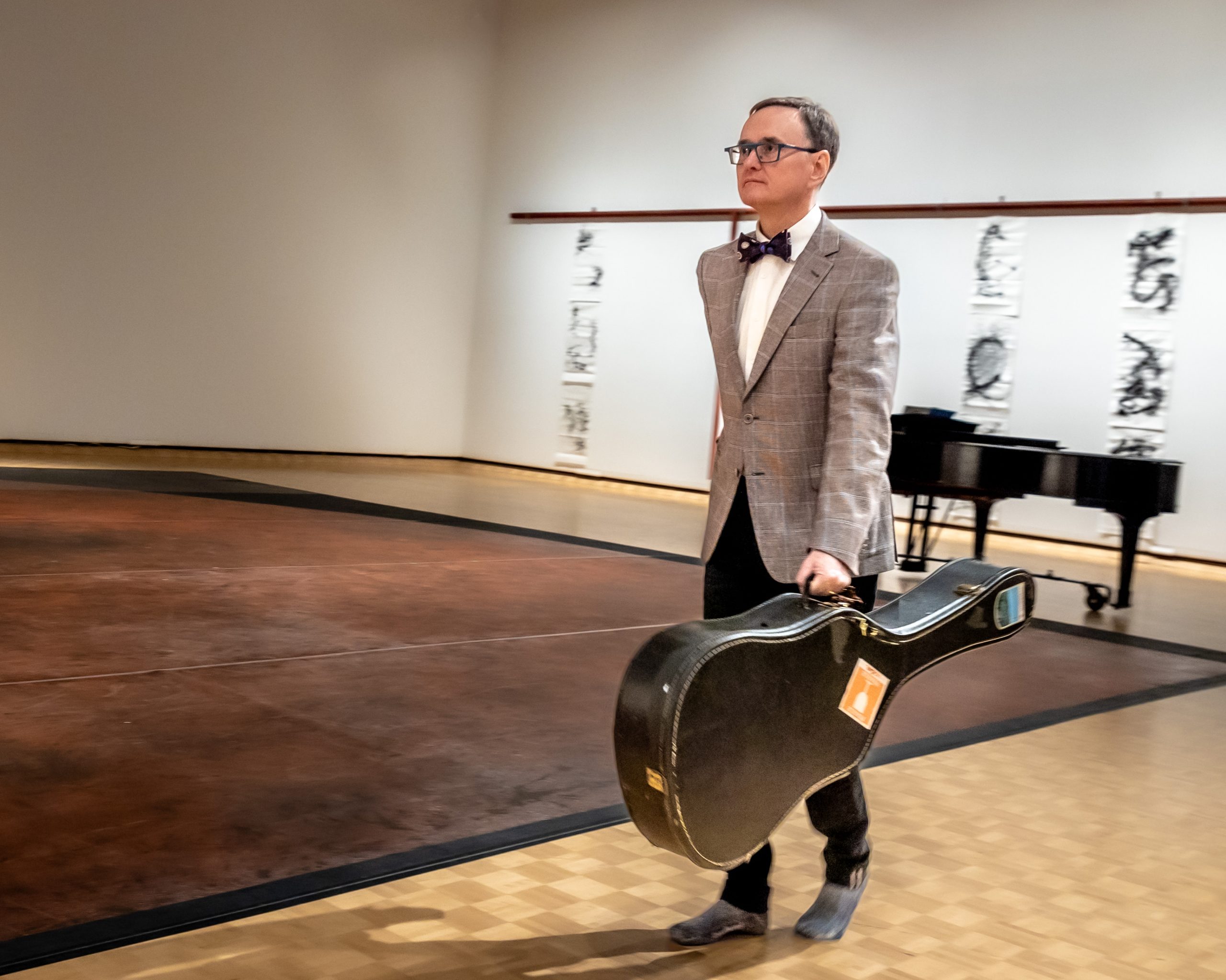 Individual in art gallery walking with guitar case.